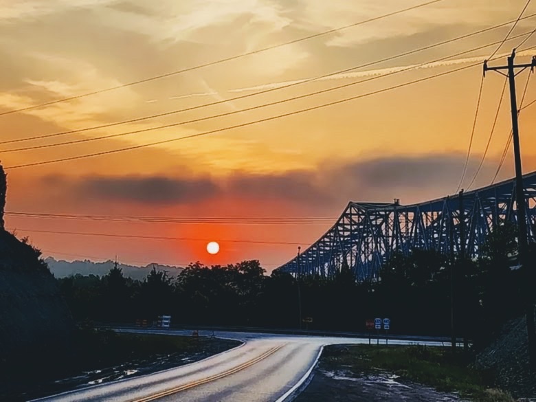 Wet highway curving toward a blue truss bridge as the sun sets