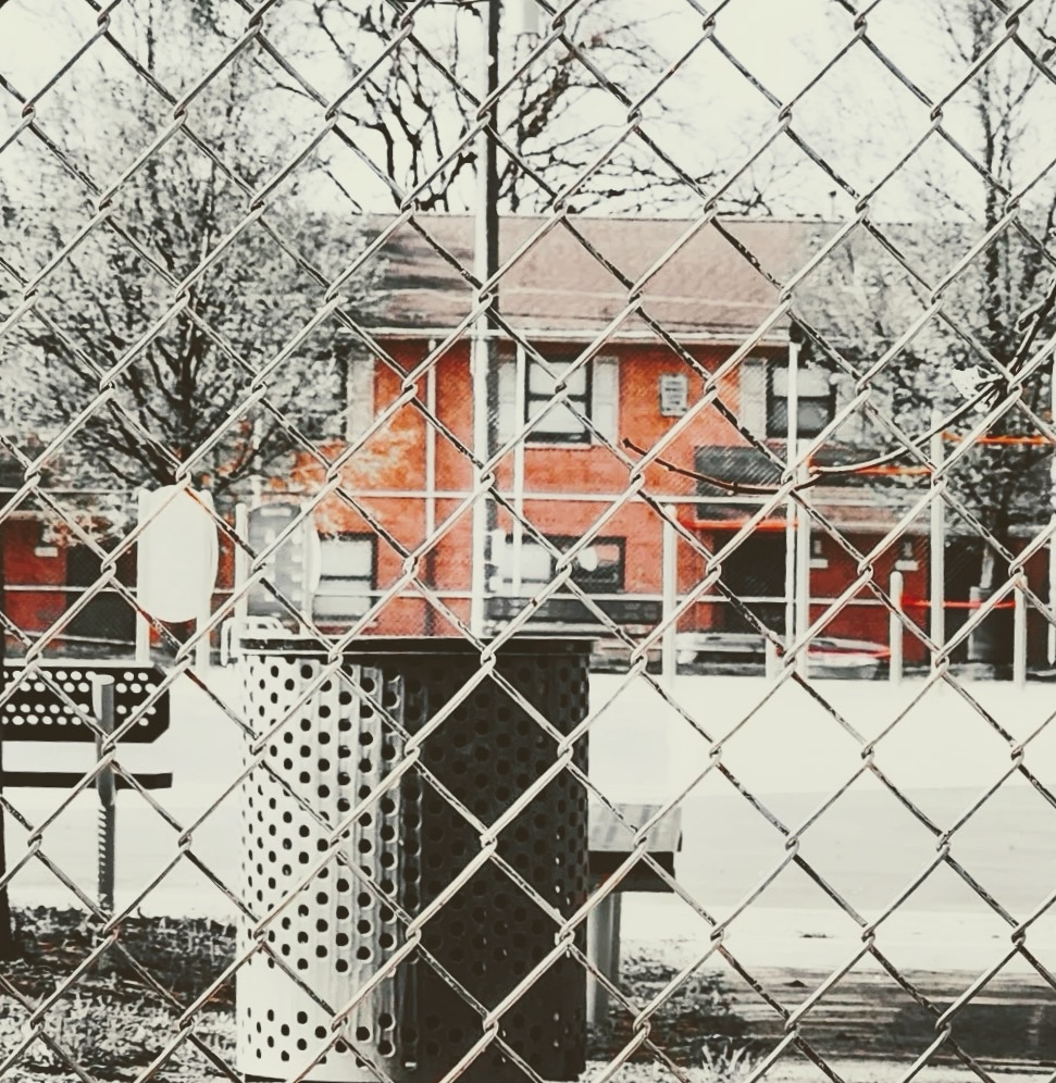 Red brick building seen through a chain link fence in near monochrome