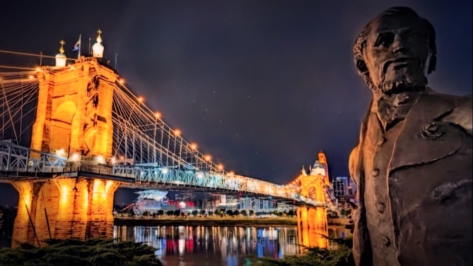 Bronze statue of John Roebling watching his lit suspension bridge at night