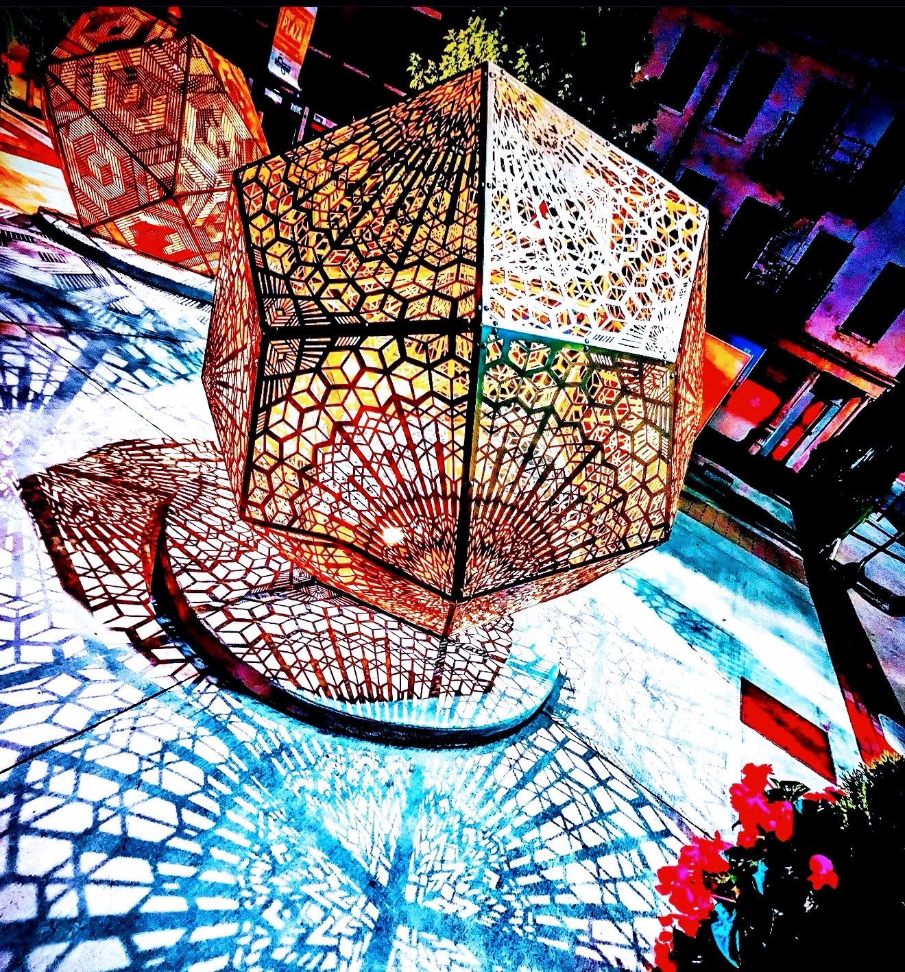 Perforated cube sculpture glowing orange and casting lace shadows on the pavement