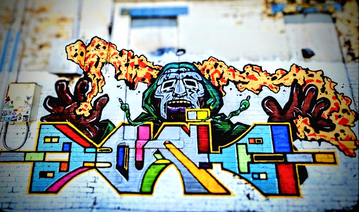 Bright graffiti piece with a masked green-hooded figure and flames on a white brick wall