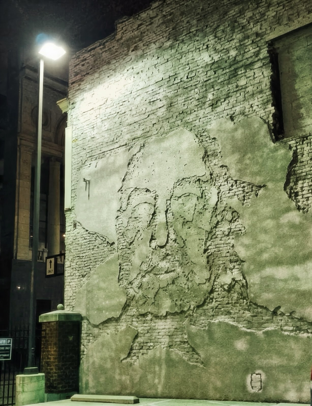 A face emerging from chipped plaster on a brick wall under a streetlight